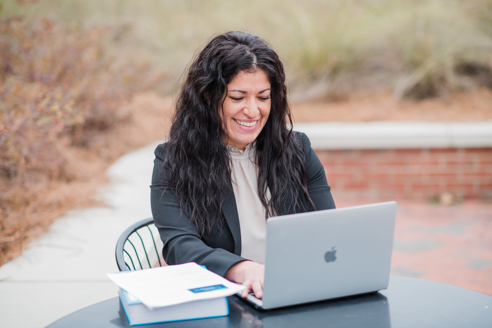 Woman in professional attire sitting at outdoor table on laptop, smiling, stack of books on left