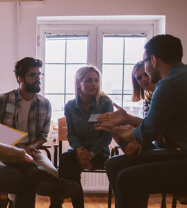 four adults sitting in a circle of chairs talking, leaning in